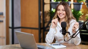Close up portrait of happy young beautiful woman in white shirt talking about different topics with followers online by laptop. Concept of interview or live broadcast for subscribers.