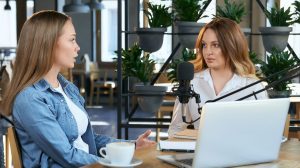 Side view of two young blogger women communicating about different topics with modern laptop and microphone. Concept of sitting in cafe and talking on broadcast by laptop for followers.